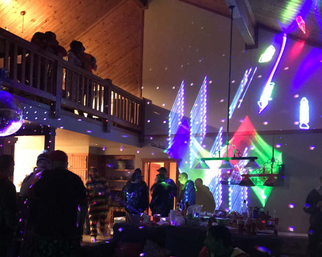 A lively indoor party scene with people gathered around, colorful lights reflecting off the walls and ceiling, and a festive atmosphere created by decorations and refreshments.