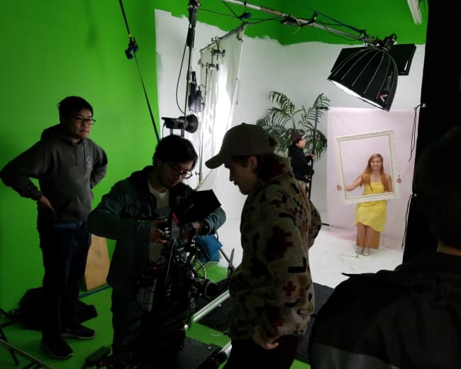 A film crew sets up equipment in a green screen studio while a girl in a yellow dress poses behind a frame.