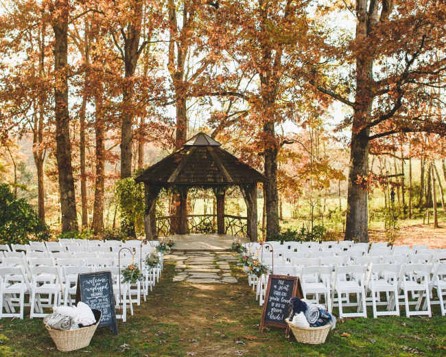 A wedding ceremony setup features rows of white chairs facing a gazebo surrounded by autumn foliage.
