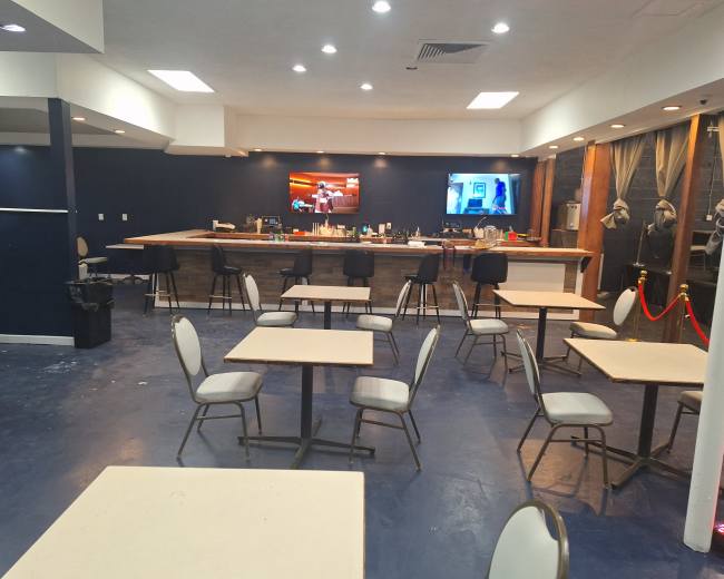The image shows a sparsely furnished room with several tables and chairs, a bar area, and two screens displaying content on the walls.