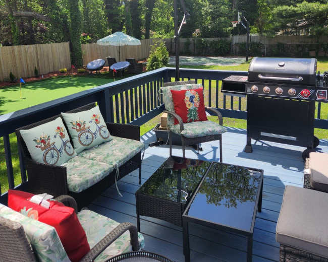 The image shows a backyard deck area with a grill, outdoor seating, and a large umbrella, surrounded by a lawn and trees.