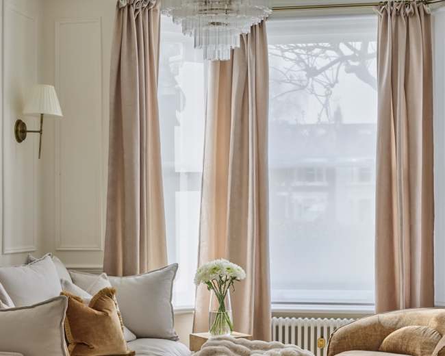 The image depicts a stylish living room with a light-colored sofa, a velvet armchair, and large windows dressed with soft curtains, accented by a textured throw blanket and a floral arrangement on a coffee table.