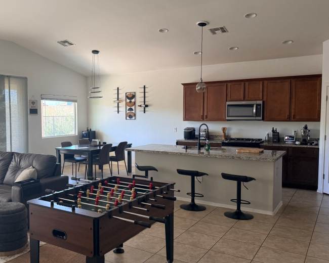 The image shows a modern living space featuring a leather sectional sofa, a foosball table, and an open kitchen with wooden cabinets and a dining area.