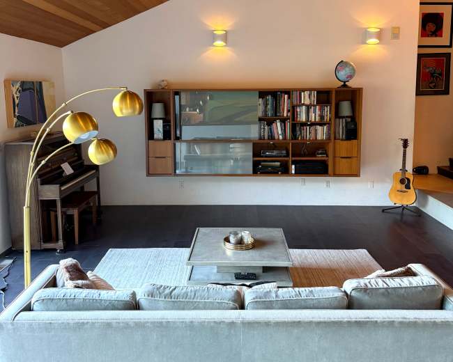 The image shows a modern living room featuring a light gray sofa, a coffee table, a wall-mounted bookshelf, and a guitar leaning against the wall.