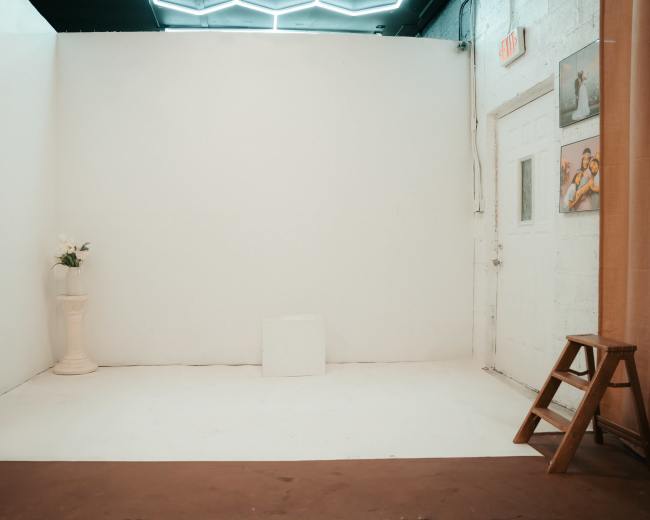 The image shows a minimalistic white room with a vase on a pedestal, a small white block on the floor, and a stool in the corner.