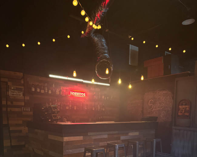 The image shows a dimly lit bar area with a wooden counter, shelves stocked with bottles, and bar stools positioned in front.