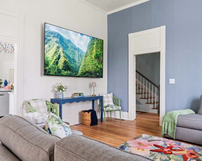 A modern living room features a large television mounted on a wall, with a view of lush mountains displayed, surrounded by comfortable seating and decor.