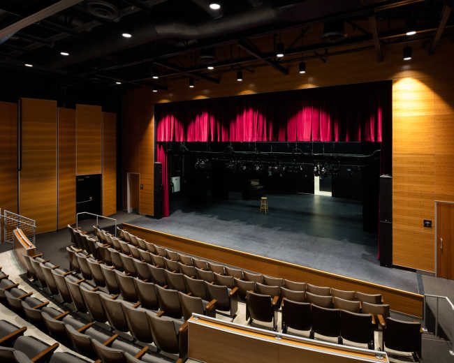 The image shows an empty theater with a stage featuring a red curtain and a single stool, surrounded by a seating area.