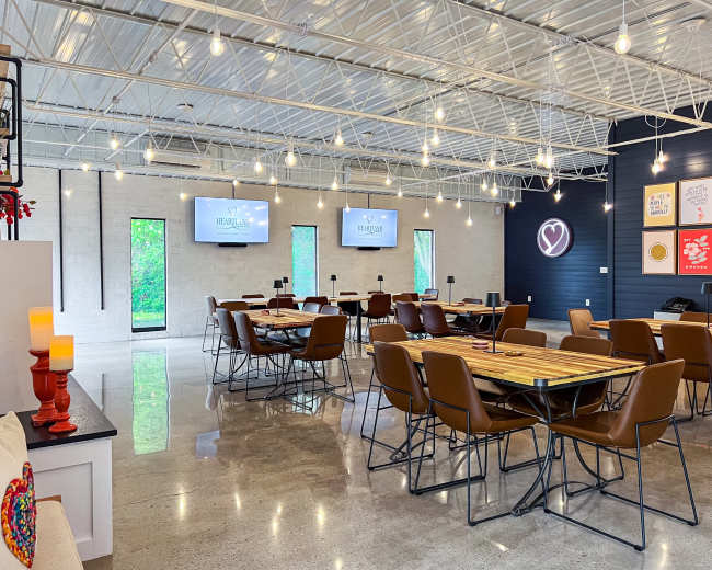 The image shows a spacious, modern dining area with wooden tables, metal chairs, and large windows letting in natural light.