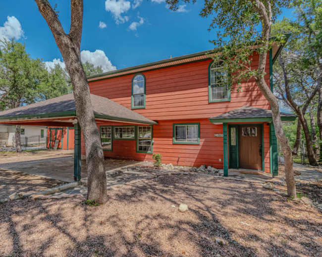 The image shows a two-story red house surrounded by trees and a gravel landscape.