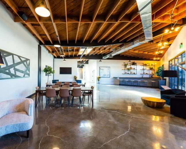 The image shows a modern interior space featuring a dining area with wooden chairs and a table, a seating area with a sofa, and a kitchenette or service area in the background, all under a wood-beamed ceiling.