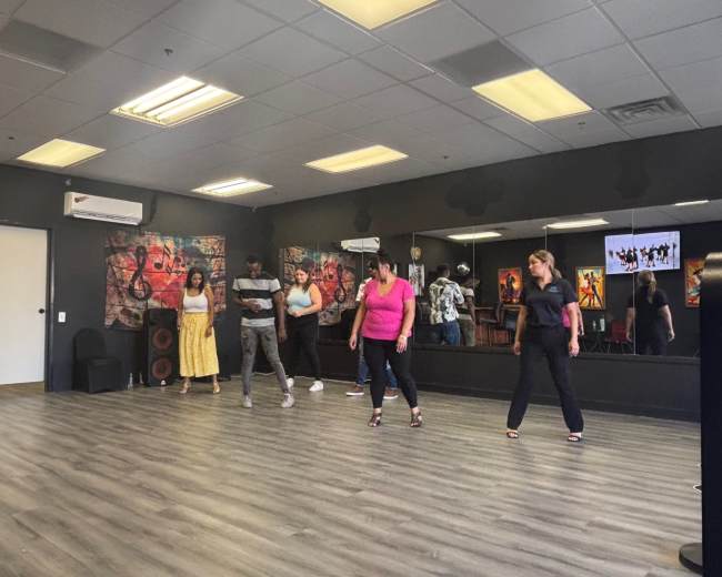 A group of six people dance in a studio with mirrors and colorful artwork on the walls.