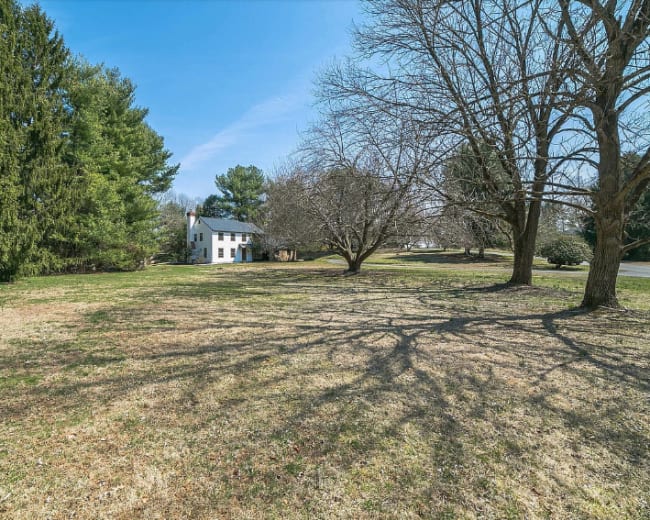 A white house stands in a grassy yard surrounded by several trees under a clear blue sky.