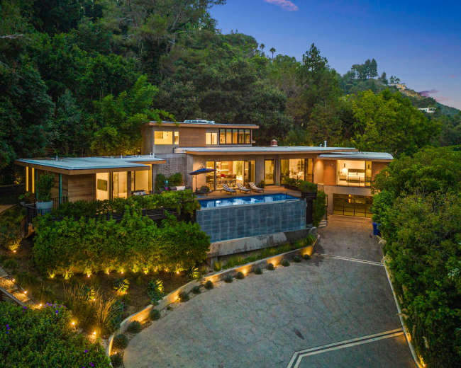 The image shows a modern, multi-level house surrounded by greenery, with a swimming pool in the backyard and a paved driveway leading up to it.