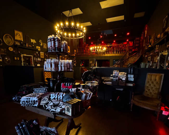 The image shows an interior of a dimly lit shop with various products displayed on tables, shelves, and a staircase that leads to an upper level.