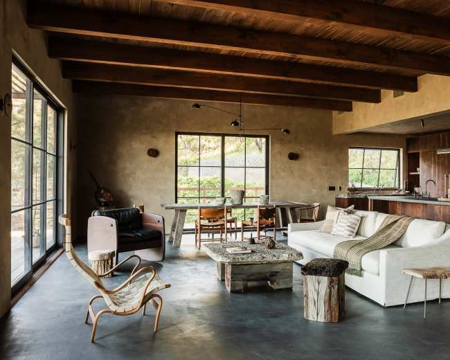 The image shows a modern living area featuring a white sofa, a wooden coffee table, and large windows that overlook a green outdoor space.