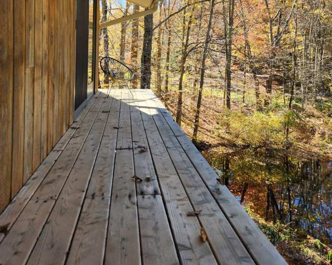 A wooden deck extends alongside a house, bordered by trees with autumn foliage and a nearby pond.
