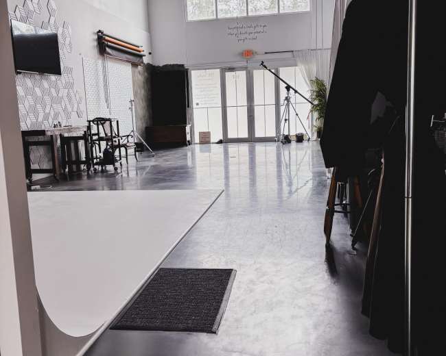 The image shows the interior of a photography studio featuring a large white backdrop, sleek concrete floors, and large windows letting in natural light.