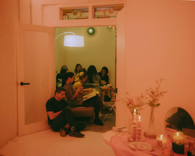 A group of people gathers around a table in a brightly lit room while another person sits on the floor near the doorway.