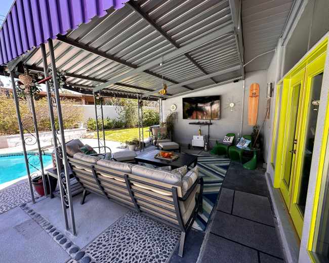 A covered outdoor patio with a seating area, a television mounted on the wall, and a view of a pool and lawn through large glass doors.