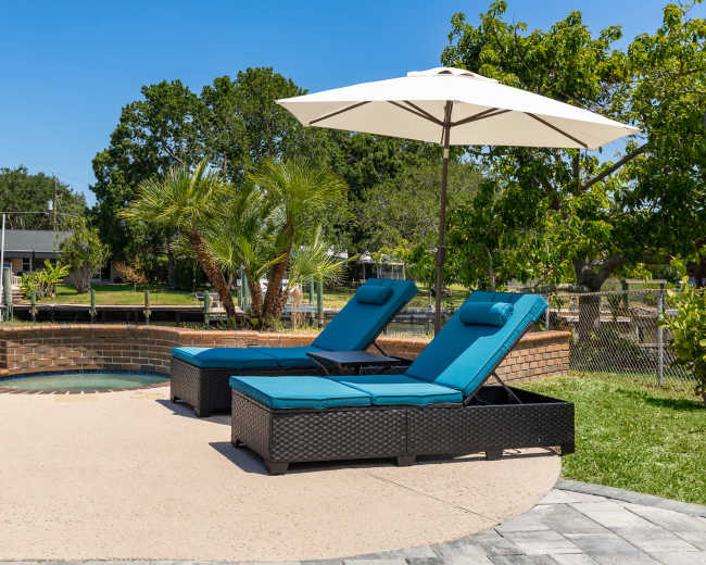 Two turquoise lounge chairs sit under a white umbrella by a pool with surrounding greenery and a low brick wall.