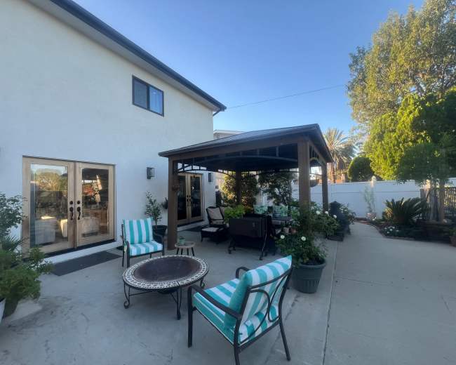 A backyard area features a covered patio with seating, a fire pit, and surrounding plants and trees.