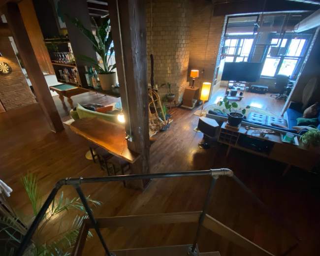 The image shows a spacious, well-lit interior featuring wood floors, a large wooden table, and various plants, along with a seating area and a television.