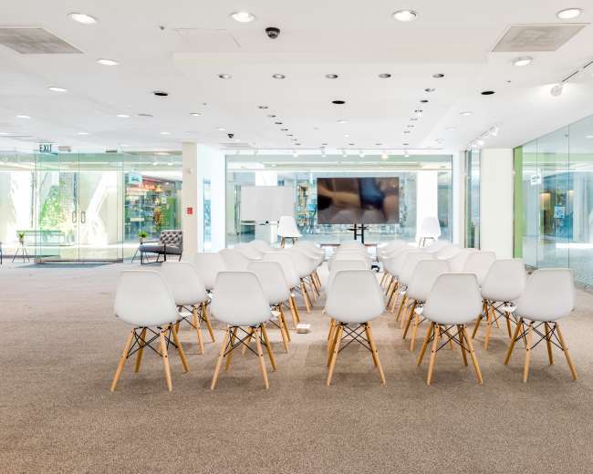 A row of white chairs with wooden legs is set up facing a large screen in a brightly lit room with glass walls.