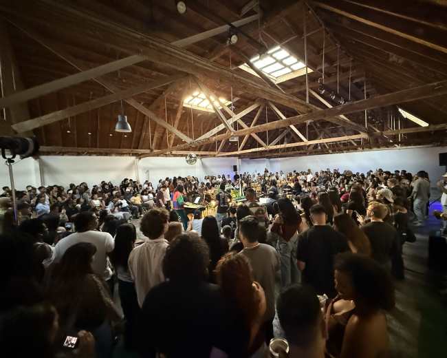 A large crowd of people gathers in an industrial space with wooden beams, watching a performance at the center.