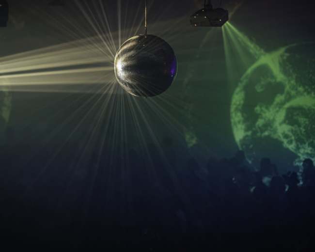 A disco ball reflects beams of light in a dimly lit venue filled with shadows and projections.
