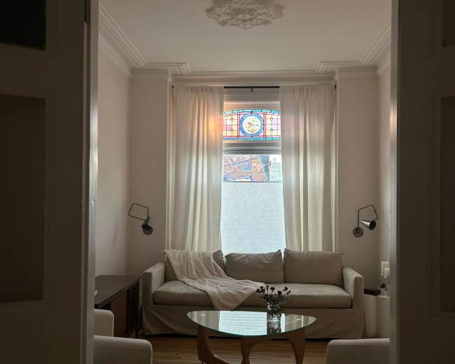 A cozy living room features a beige sofa, two armchairs, a glass coffee table, and a stained glass window.