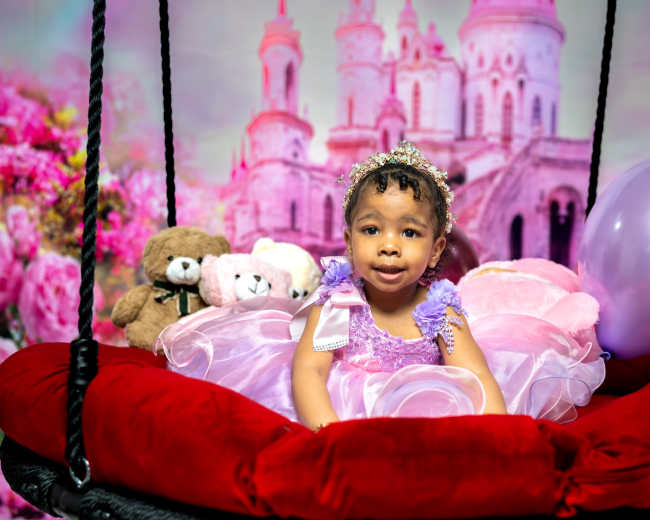 A young girl in a colorful dress sits on a swing surrounded by plush toys and balloons, with a castle backdrop.