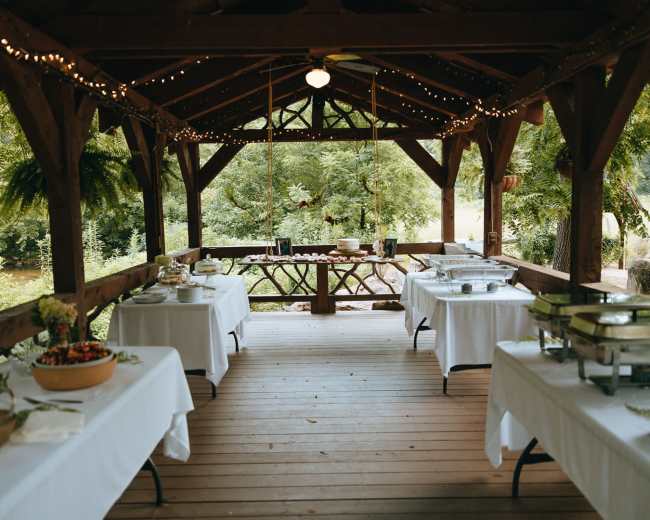A wooden pavilion is set up with several tables covered in white tablecloths, displaying food and decorations surrounded by greenery.