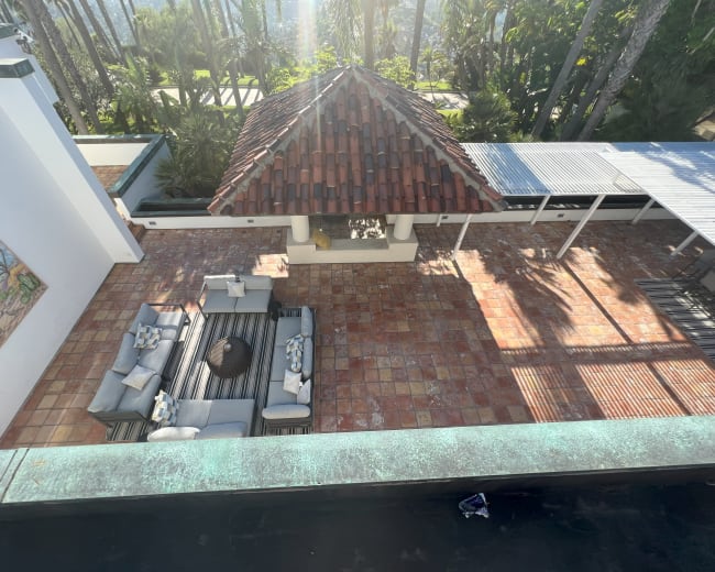 The image shows a spacious outdoor patio area with a tiled floor, a covered seating arrangement, and a traditional tiled roof structure surrounded by palm trees.