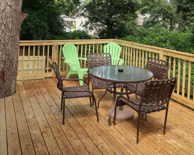 A wooden deck features a round glass table surrounded by six chairs, with a large tree providing shade overhead.