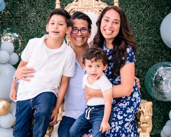 A family of four, including two boys and their parents, posing together on an ornate chair in front of a decorative backdrop with balloons.