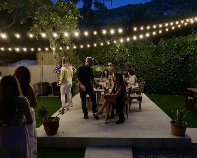 A group of people socialize in a backyard setting under string lights, surrounded by greenery and potted plants.