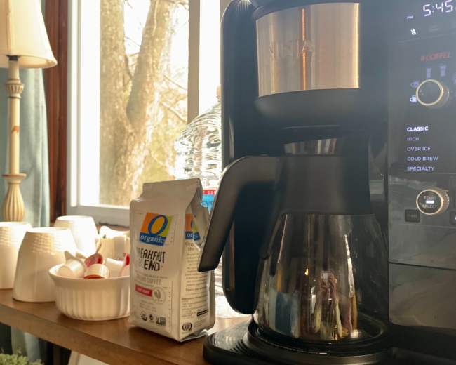 A coffee maker sits on a table next to a bag of coffee grounds and several cups, with sunlight streaming through nearby windows.