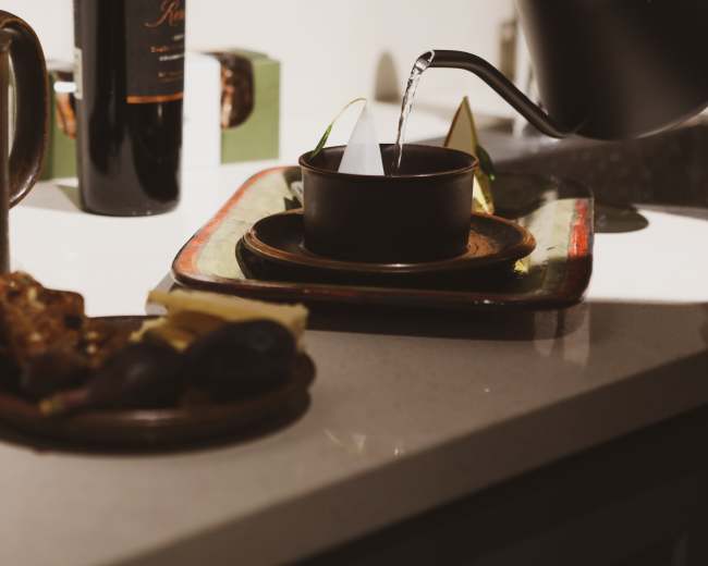 A teapot pours water into a dark ceramic cup placed on a decorative tray, alongside a plate of assorted fruits and snacks.