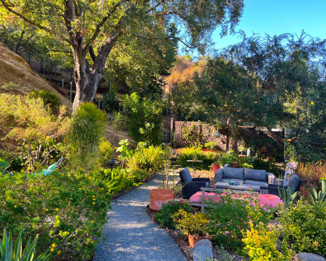 A winding gravel path leads through a vibrant garden filled with various plants and comfortable seating areas under a clear blue sky.