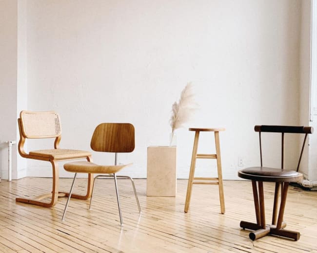 The image shows four different styles of chairs arranged in a minimalist space with a light-colored wall and a simple pedestal displaying a decorative plant.