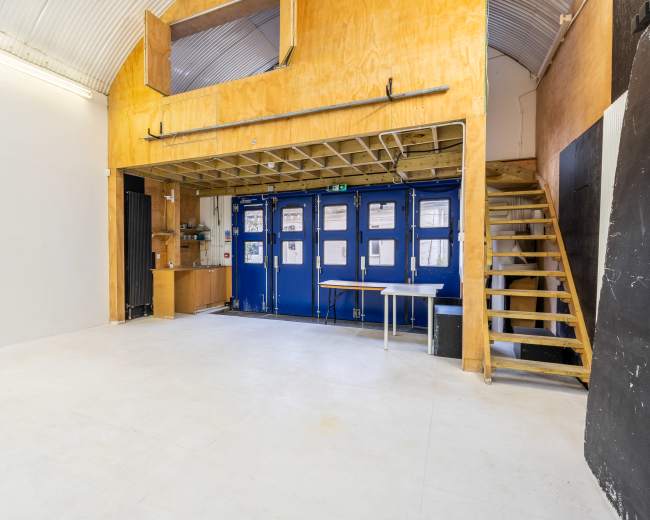 The image shows an empty interior space with a wooden loft area, blue doors, a staircase, and a table in the foreground.