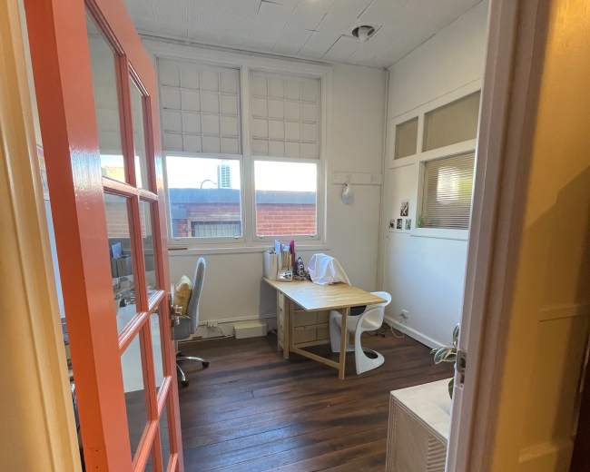 The image shows a narrow office space with wooden floors, a desk and chair, and large windows with blinds.