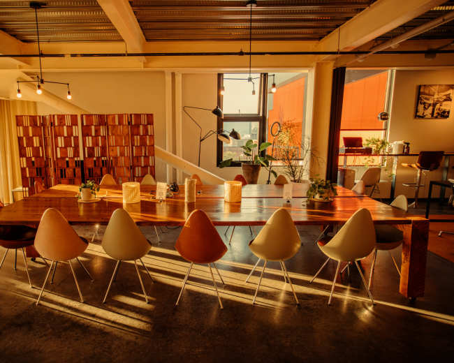 A long wooden table with a row of colorful chairs is set in a brightly lit room featuring large windows and plants.