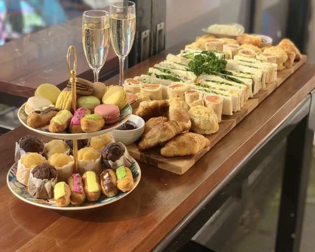 The image shows a table displaying an assortment of sandwiches, pastries, and desserts, accompanied by two glasses of champagne.