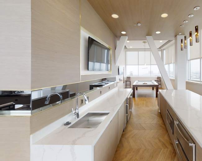 The image shows a modern kitchen with a long island counter, sleek cabinetry, and large windows offering city views.