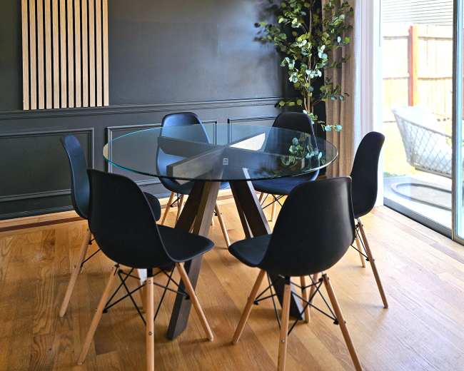 The image shows a modern dining area with a glass table surrounded by black chairs and a decorative light fixture above, complemented by indoor plants and wooden accents on the wall.