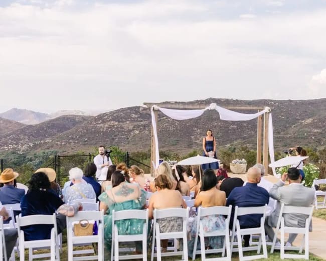 A wedding ceremony takes place outdoors with guests seated at tables, and a couple standing under a decorated arbor against a mountainous backdrop.