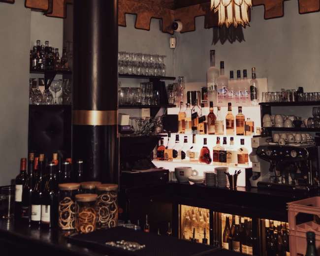 The image shows a bar area featuring shelves stocked with various bottles, a coffee machine, and decorative lighting under a patterned ceiling.