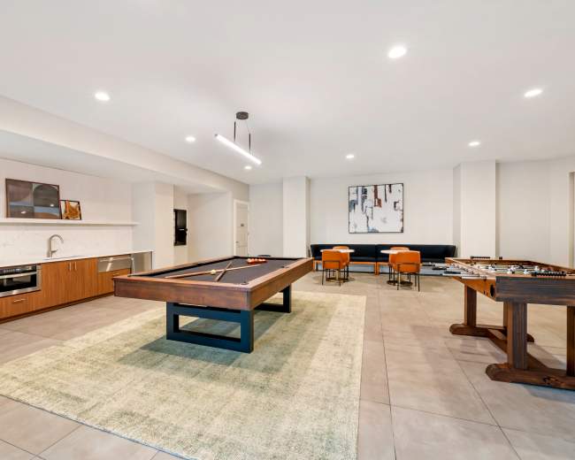 The image shows a spacious recreational room featuring a pool table, foosball table, kitchen area, and seating arranged on a plush rug.
